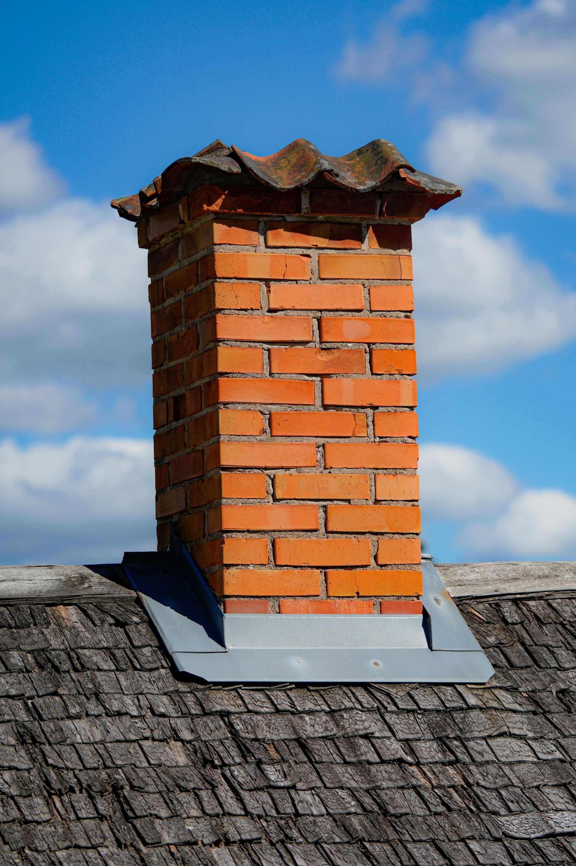 A red brick chimney with a weathered top cap, surrounded by lead flashing on a textured gray shingled roof against blue.