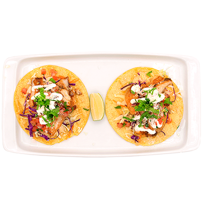 Two tacos on a rectangular white plate with lime wedge; topped with meat, vegetables, and sauce.