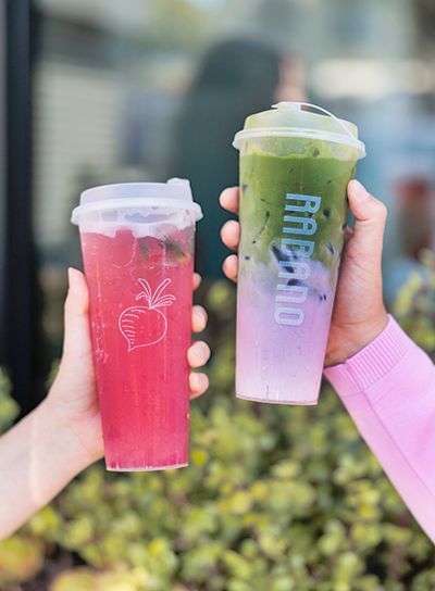 Two hands holding colorful iced drinks in plastic cups, one pink, one layered purple and green.