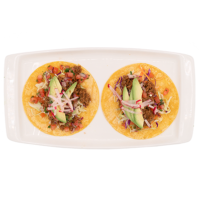 Two tacos on a white rectangular plate. They are topped with meat, avocado, and vegetables.