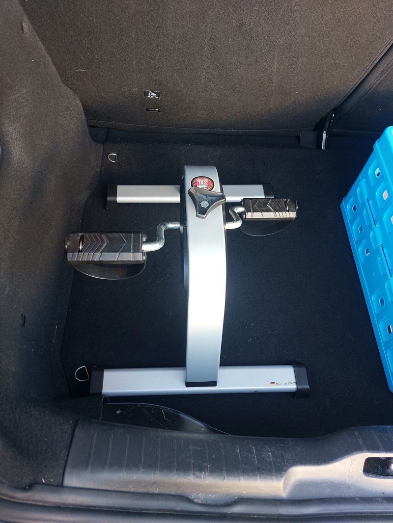 Een zilverkleurig voetpedaalapparaat ligt in de kofferbak van een auto, centraal op het zwarte tapijt.