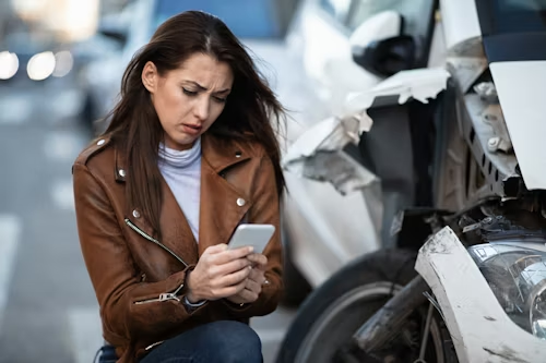 Woman in a brown jacket using a phone beside a damaged car after a collision