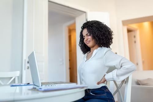 Woman at a desk with laptop, grimacing and clutching her lower back.