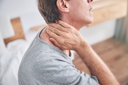 Man holding his neck, grimacing. Wearing gray shirt, indoors.