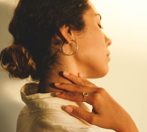 A person in profile view with hair in a bun, wearing a hoop earring and a ring, hand gently touching their neck.