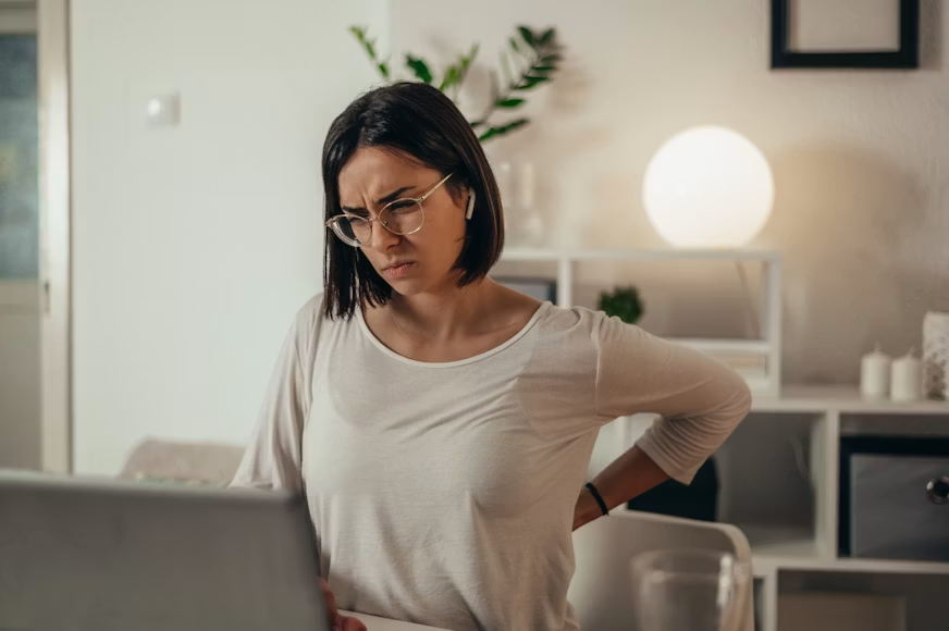 Woman in white shirt rubbing her lower back while working at a laptop in a home office