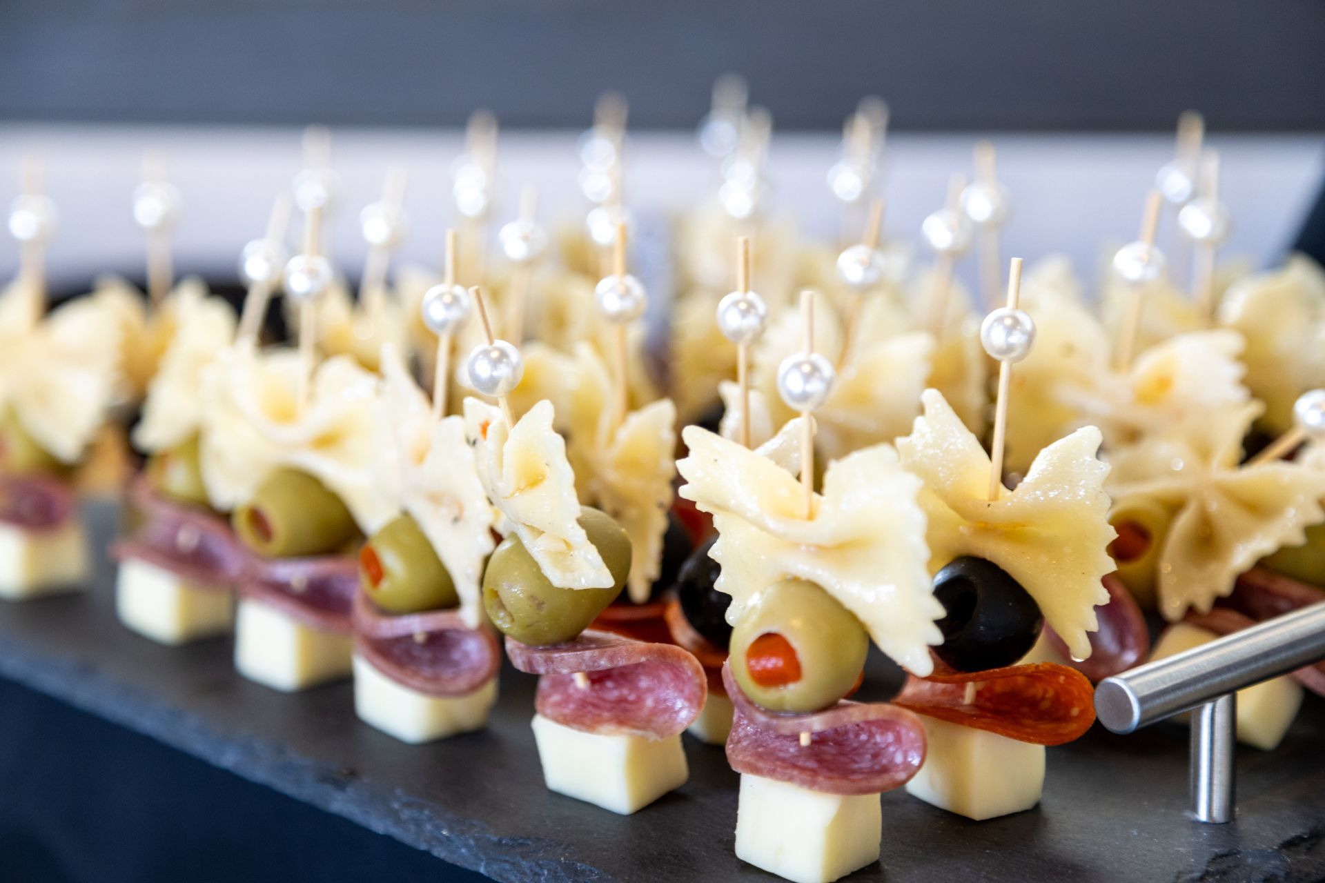 Appetizer skewers: cheese cubes, salami, olives, pasta, and pearl-topped toothpicks on a slate platter.