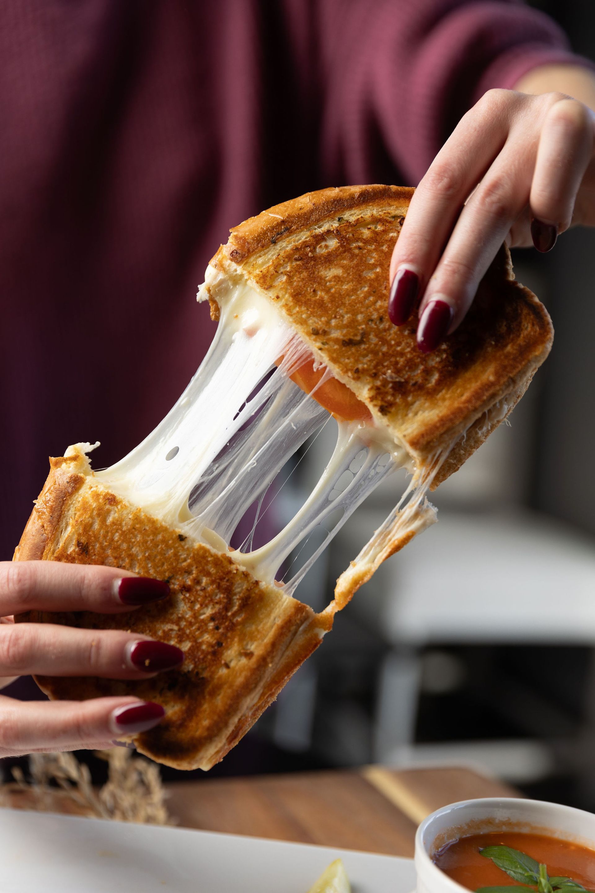 Person holding a grilled cheese sandwich, with melted cheese stretching; tomato soup bowl in foreground.