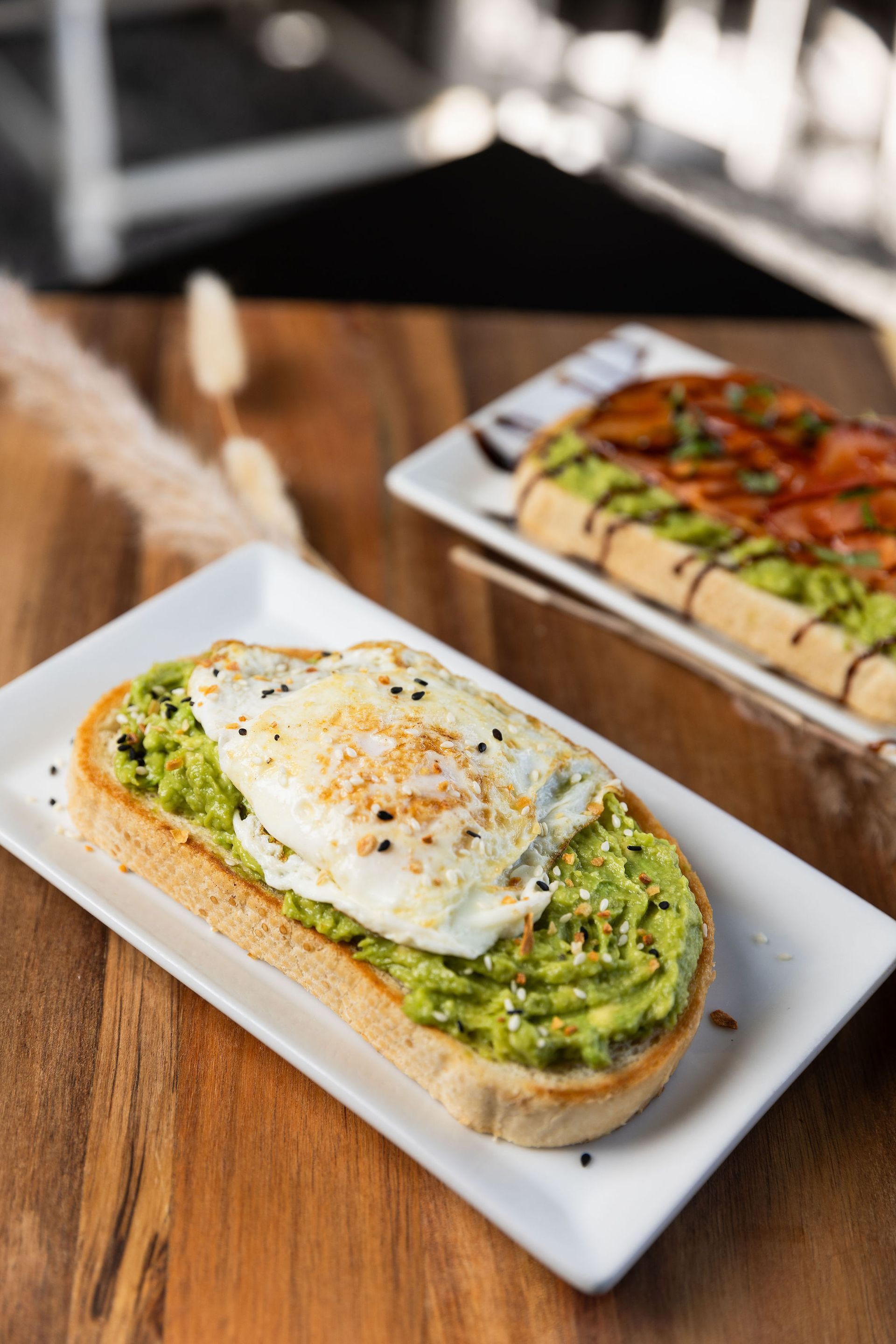 Avocado toast with fried egg on white plate, another with toppings and sauce in background, on wood table.