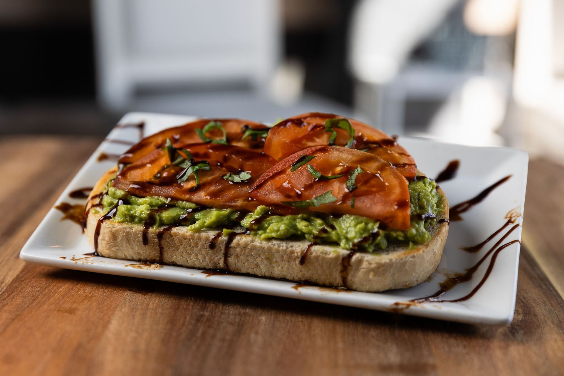 Avocado toast with tomato slices drizzled with balsamic glaze, on a white plate.