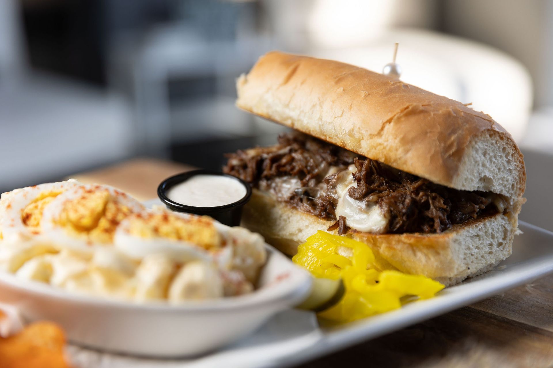 Philly cheesesteak sandwich with coleslaw, pickles, and dipping sauce on a white plate.