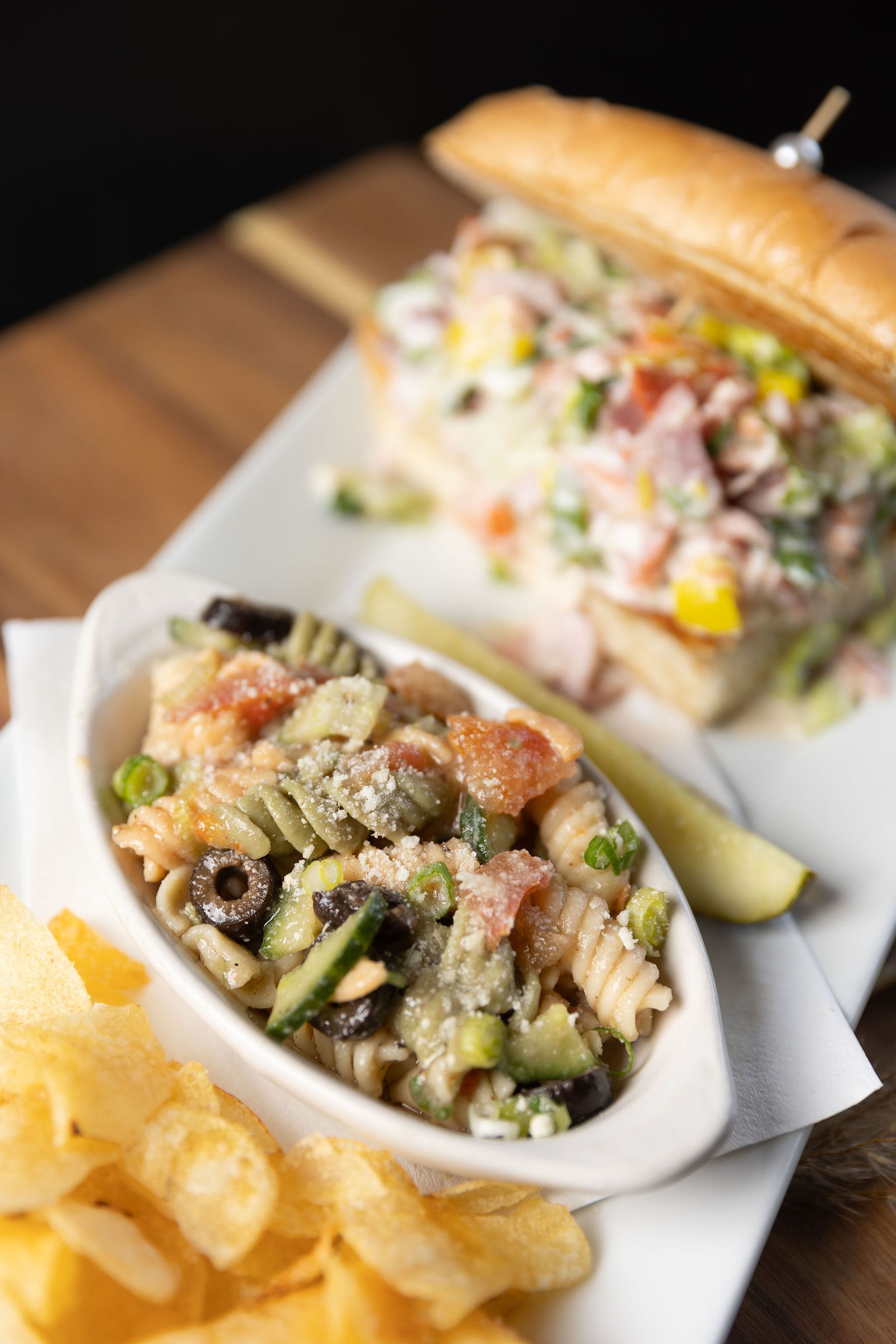 Sub sandwich with pasta salad, pickles, and chips on a wooden surface.