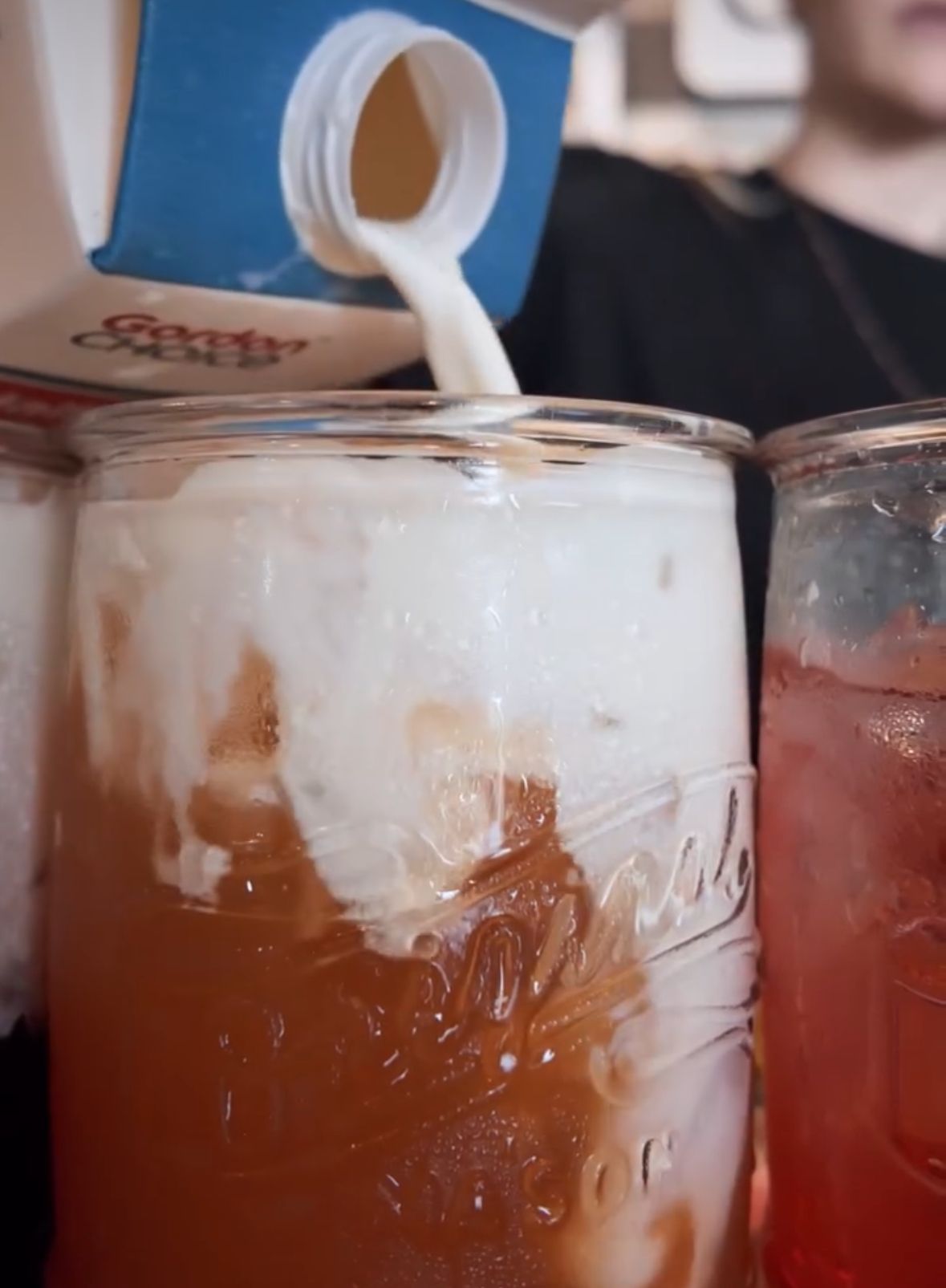 A clear glass mug overflowing with whipped cream, sprinkles, and a drink, in front of a brick wall, and a white flower.