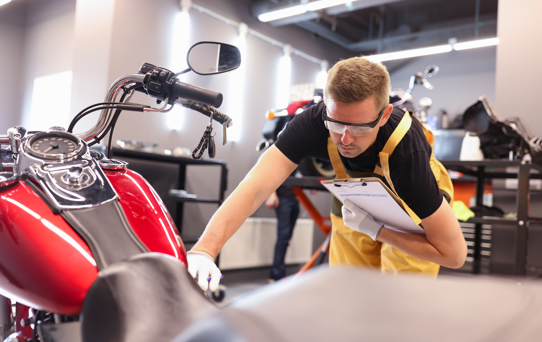 Mechanic inspecting a red motorcycle in a shop, wearing safety glasses and a clipboard.