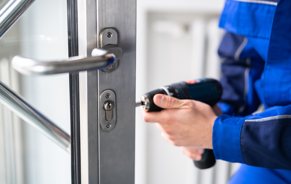 Person in blue overalls using a drill to install or repair a door lock on a metal door.