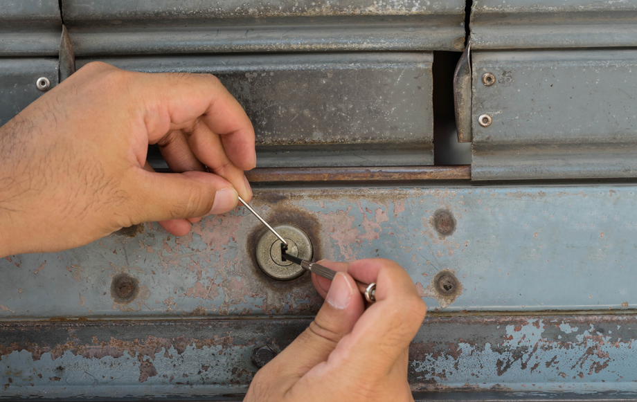 Hands picking a lock on a rusty metal door using a tension wrench and lock pick.