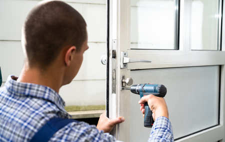 A person in a blue plaid shirt using a power drill on a door lock.