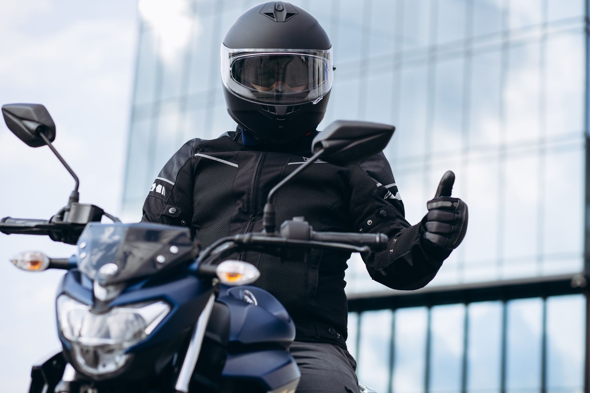 Motorcyclist in black helmet and jacket on a blue motorcycle, giving a thumbs-up gesture.