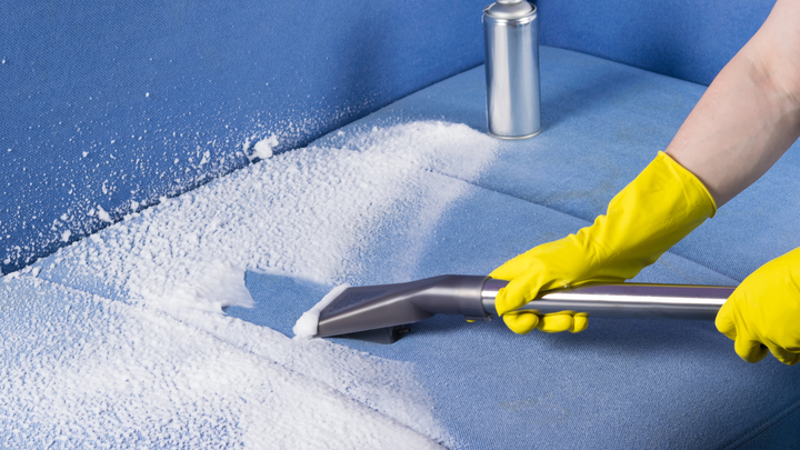 A person wearing yellow gloves is cleaning a blue couch with a vacuum cleaner.