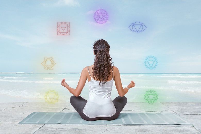 A woman is sitting on a yoga mat on the beach with chakras surrounding her.
