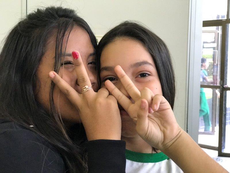 Two young girl students smiling and making the peace sign with their hands, covering their faces. 