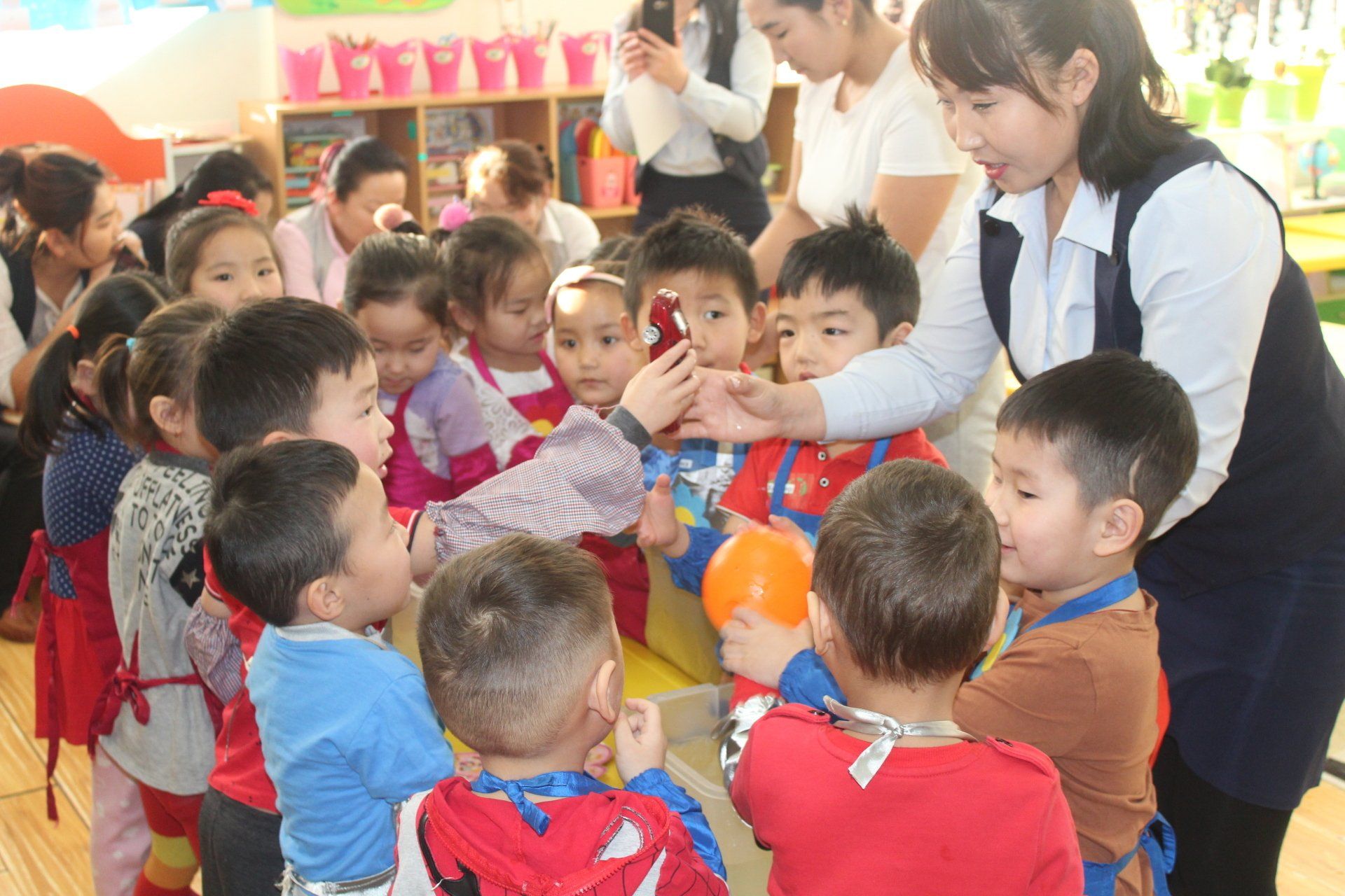 A group of young students gathered around a teacher; the teacher is handing a toy model car to one of the students. 