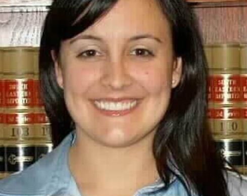 A woman is smiling in front of a shelf of books.