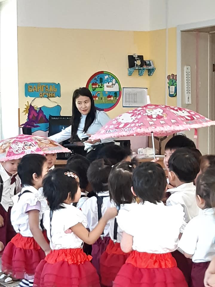 A group of students in a classroom with their teacher, gathered around holding 'Hello Kitty' umbrellas for a learning activity. 