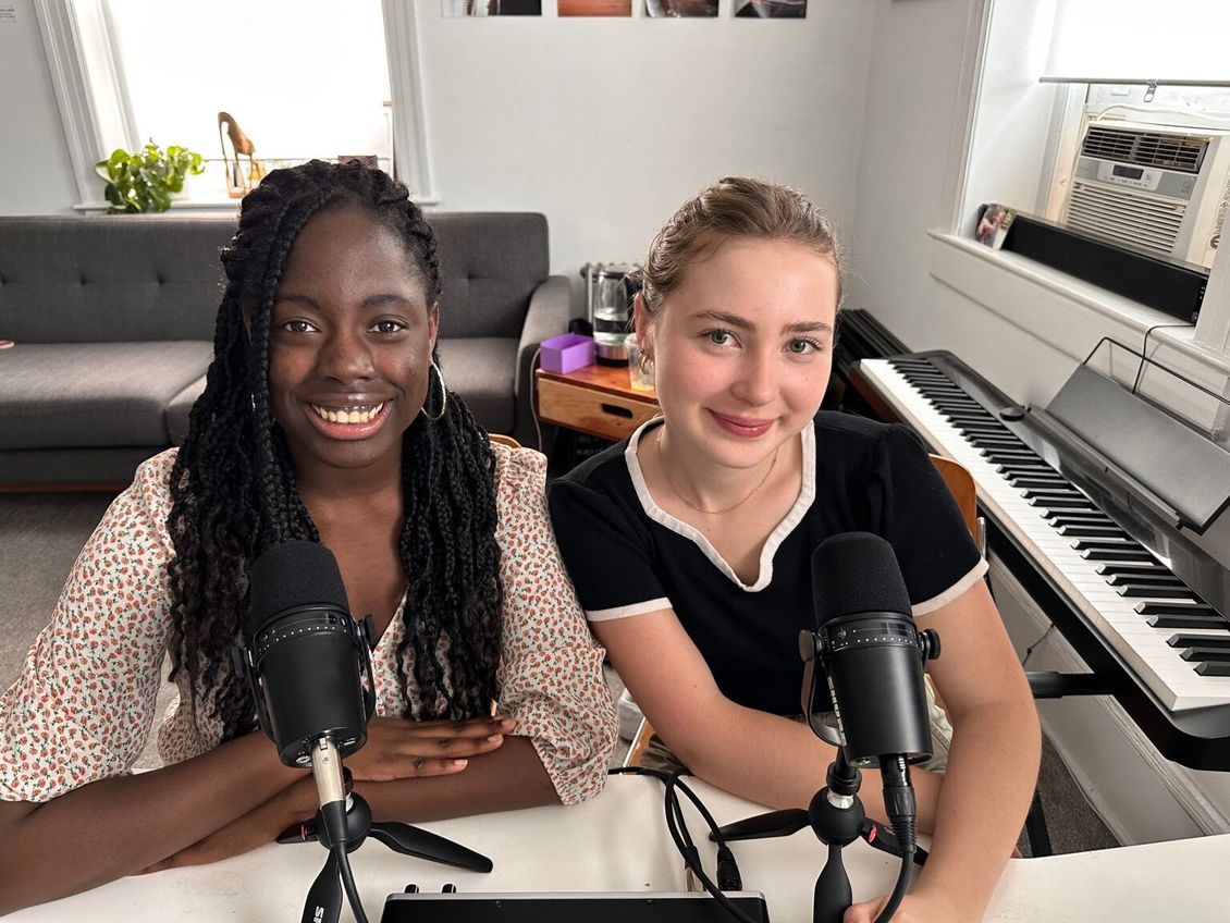Two women are sitting at a table with microphones and a piano.