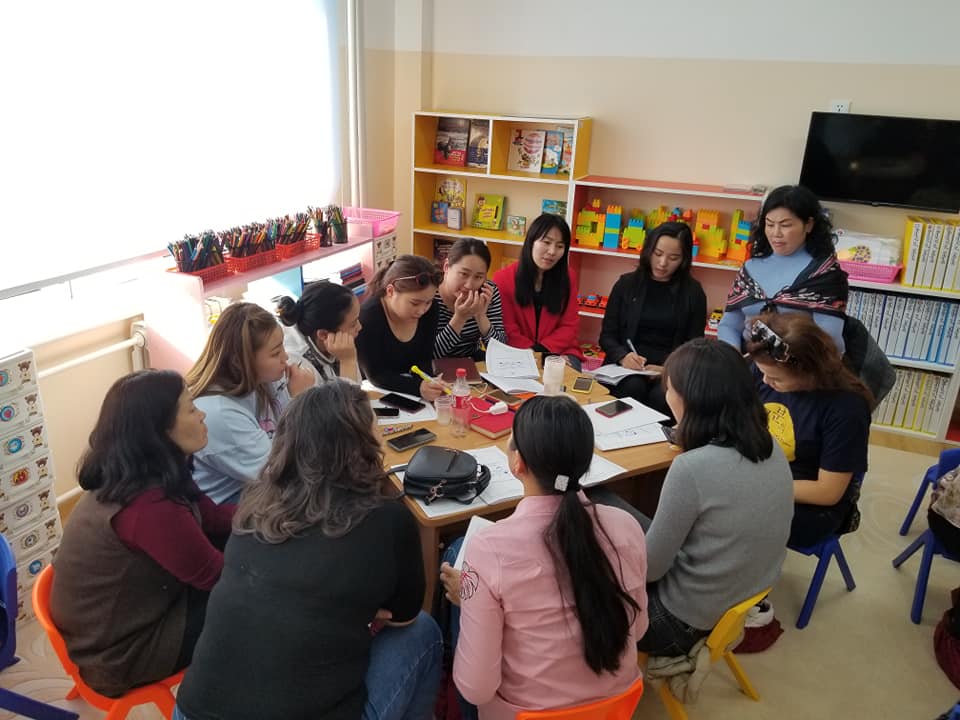A group of parents gathered around a table in a classroom participating in a facilitated conversation with student teachers. 