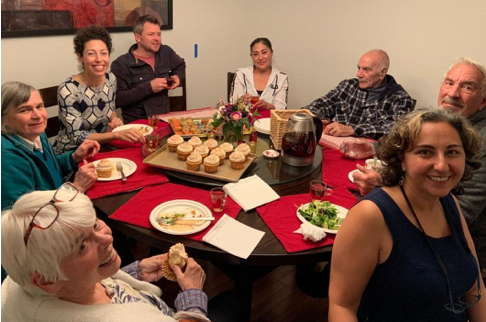 A small gathering of 8 people in a home, sitting around a table with food and beverages discussing Mona's mission.