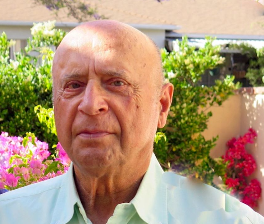 A bald man in a green shirt is standing in front of a bush with pink flowers.
