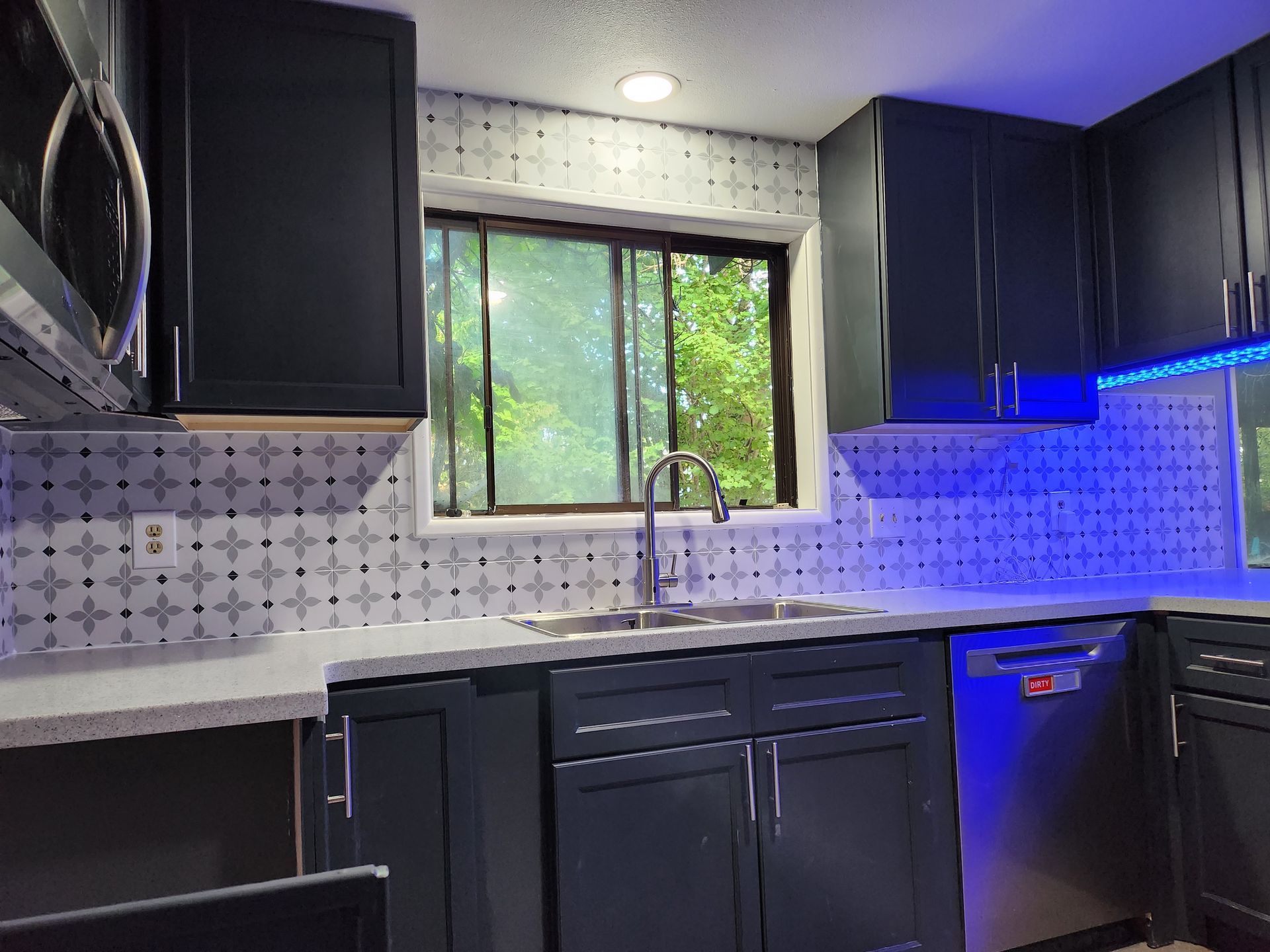 A kitchen with black cabinets , stainless steel appliances , a sink , and a window.