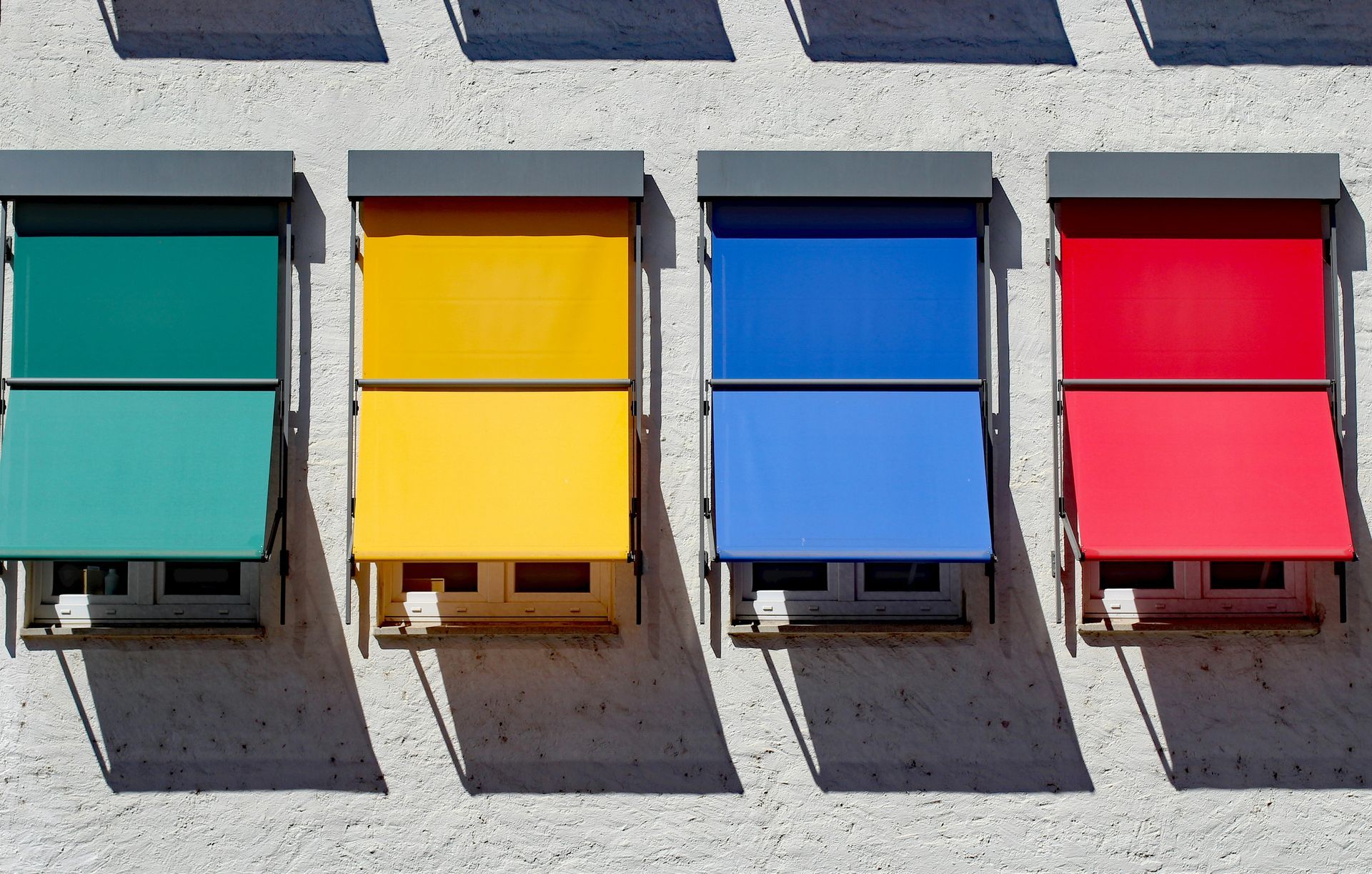 Four rectangular, colorful awnings in teal, yellow, blue, and red project from a textured wall, casting long shadows.