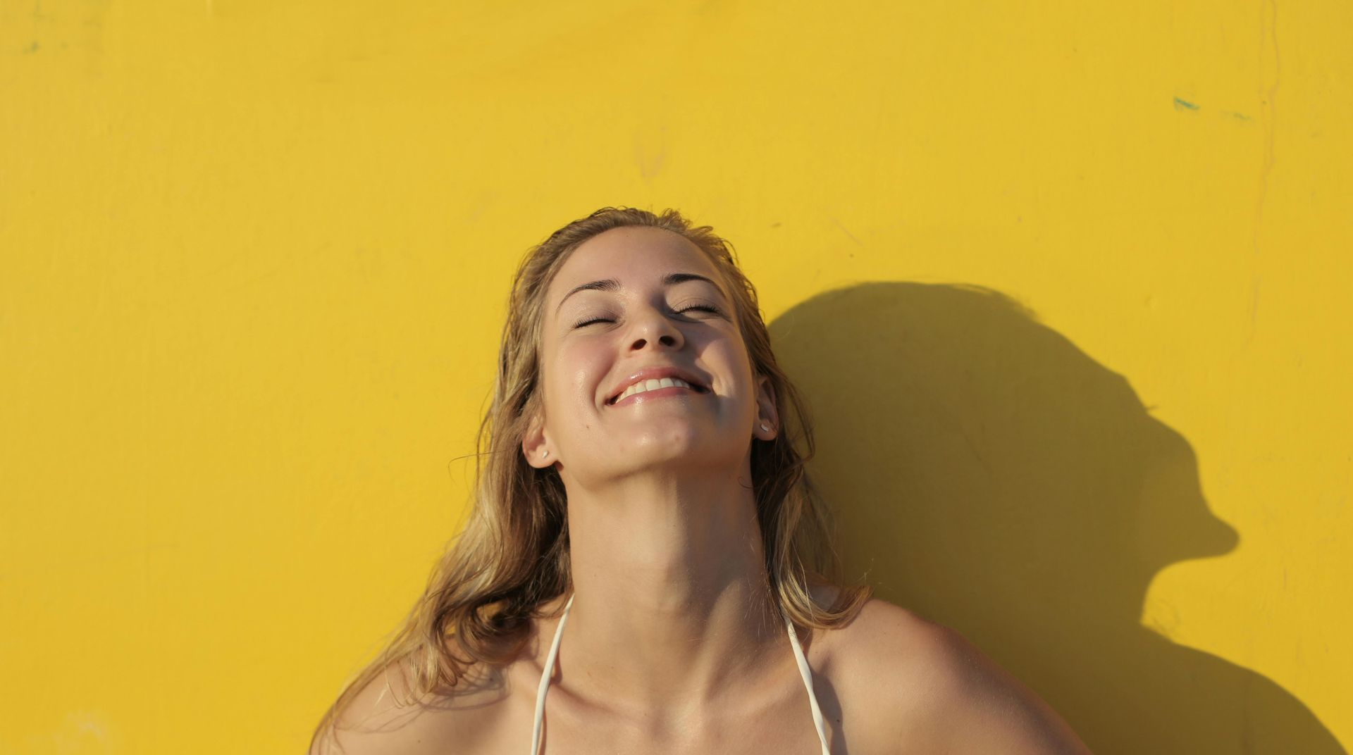 Eine Person mit lächelndem Gesichtsausdruck lehnt sich im Sonnenlicht an eine hellgelbe Wand und wirft einen Schatten nach rechts.