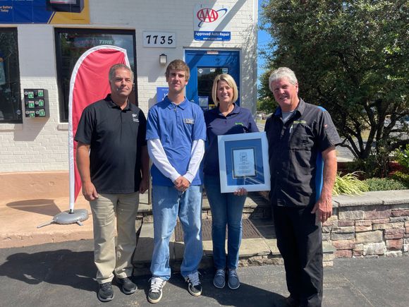 Four people standing outside an auto shop, holding a framed certificate.
