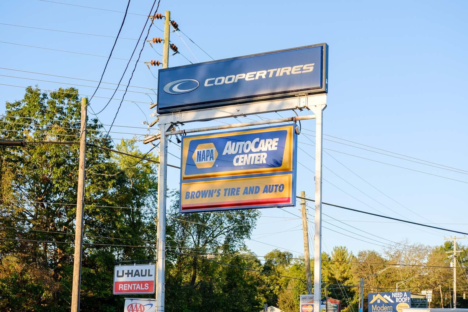 Sign for Cooper Tire's and Napa Auto Care Center with a U-Haul sign beneath, trees and power lines in the background.