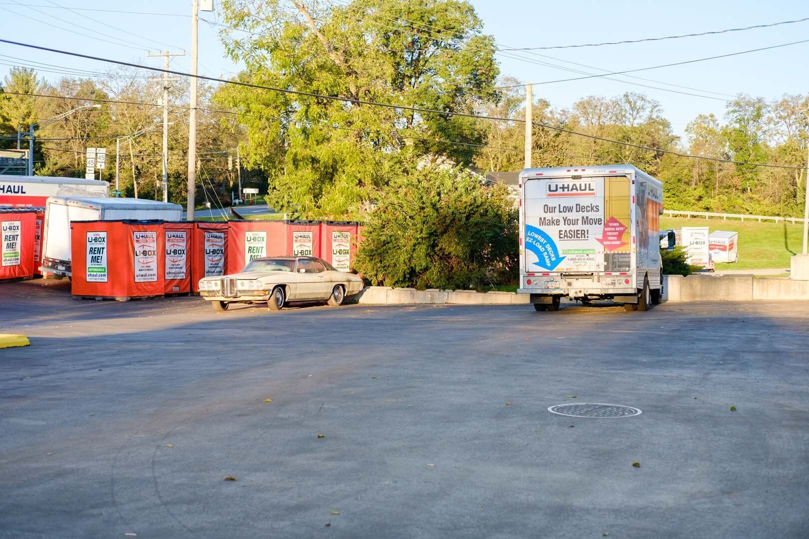 Parking lot with red storage containers, a vintage car, and a delivery truck. Trees and power lines in the background.