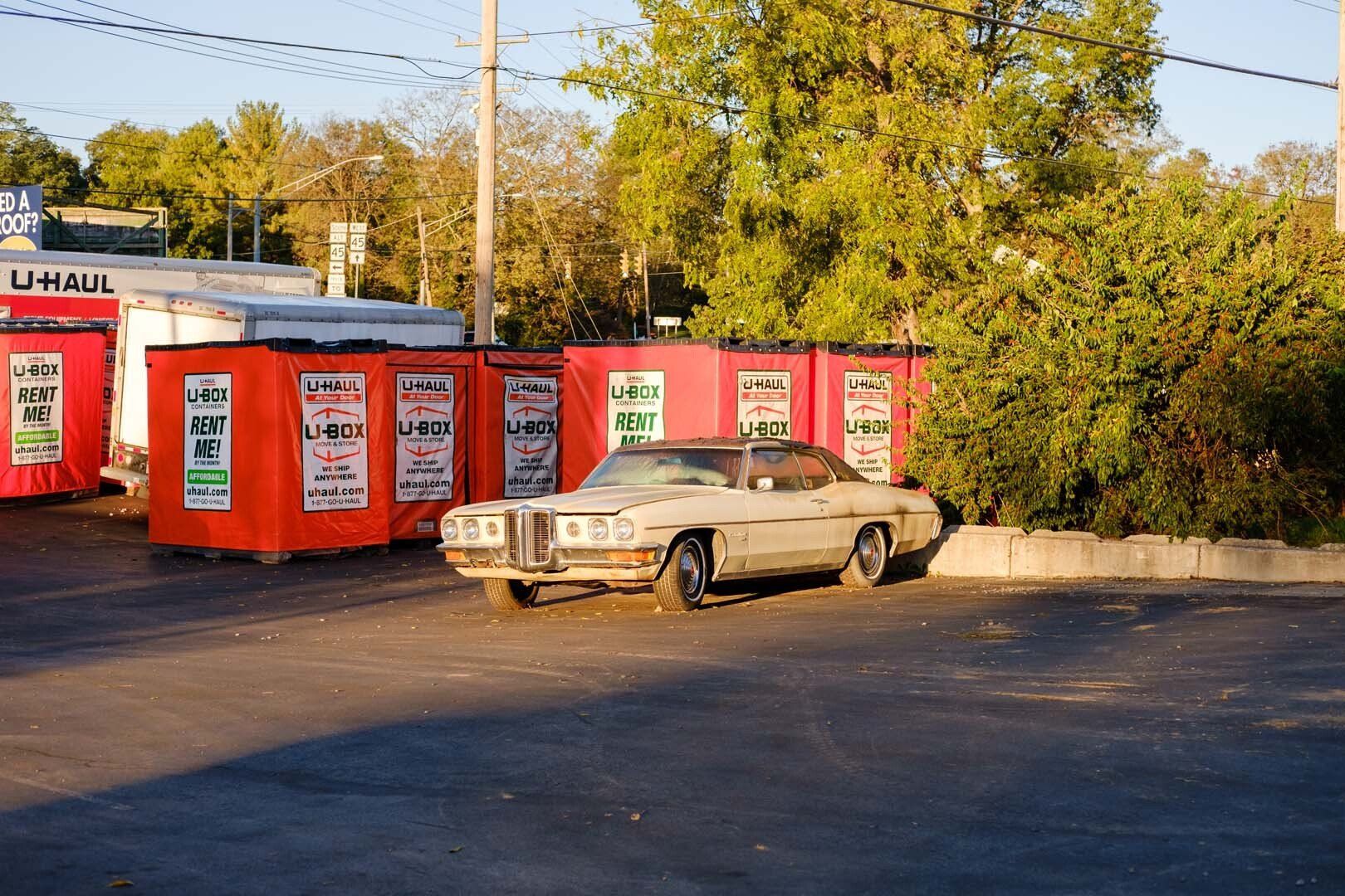 Old, beige car parked in front of red storage units. Pavement in foreground with trees and building in the background.