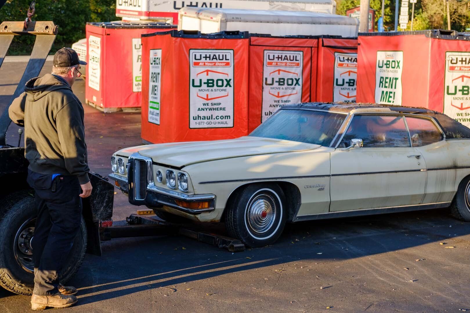 Man near a beige classic car being towed, U-Haul storage containers in background.