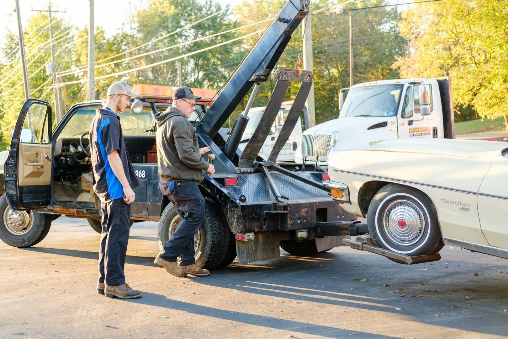 Two men loading a cream-colored car onto a tow truck outdoors.
