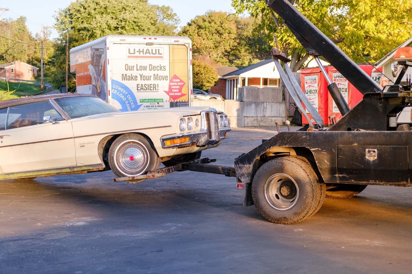 Tow truck towing a beige car on a street, a U-Haul truck and houses in the background.