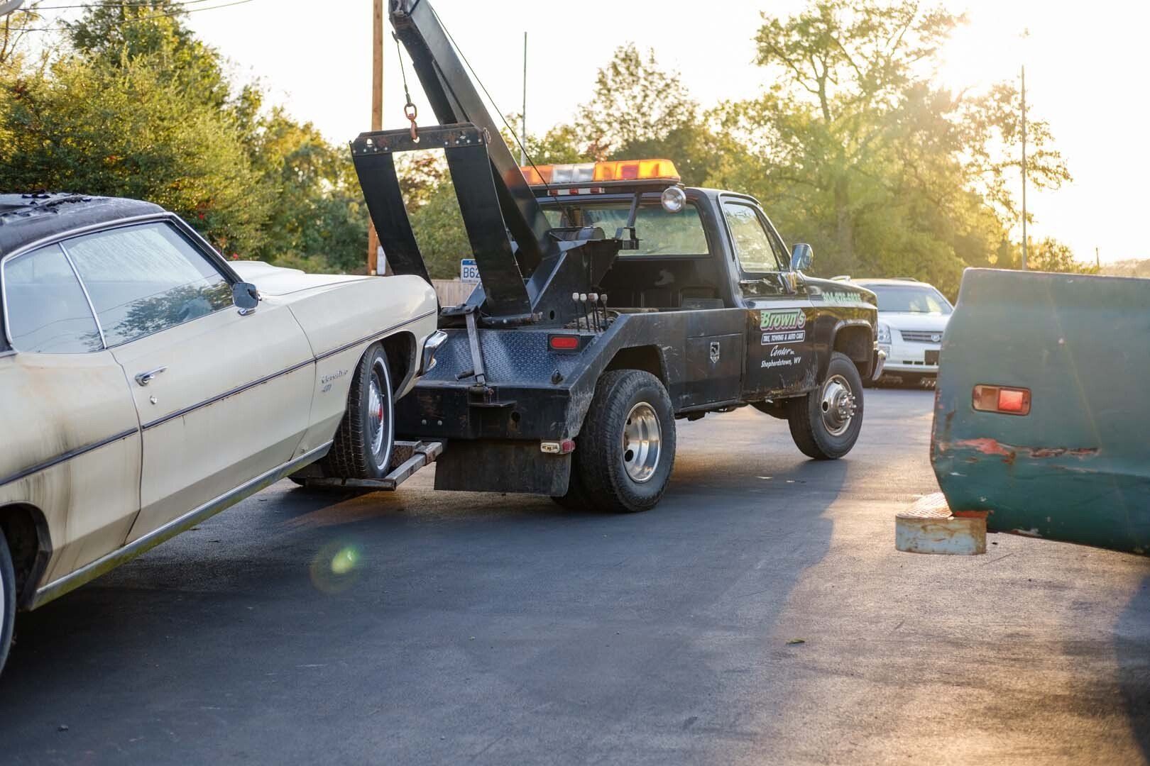 Tow truck towing a beige classic car in a parking lot. Setting sun.