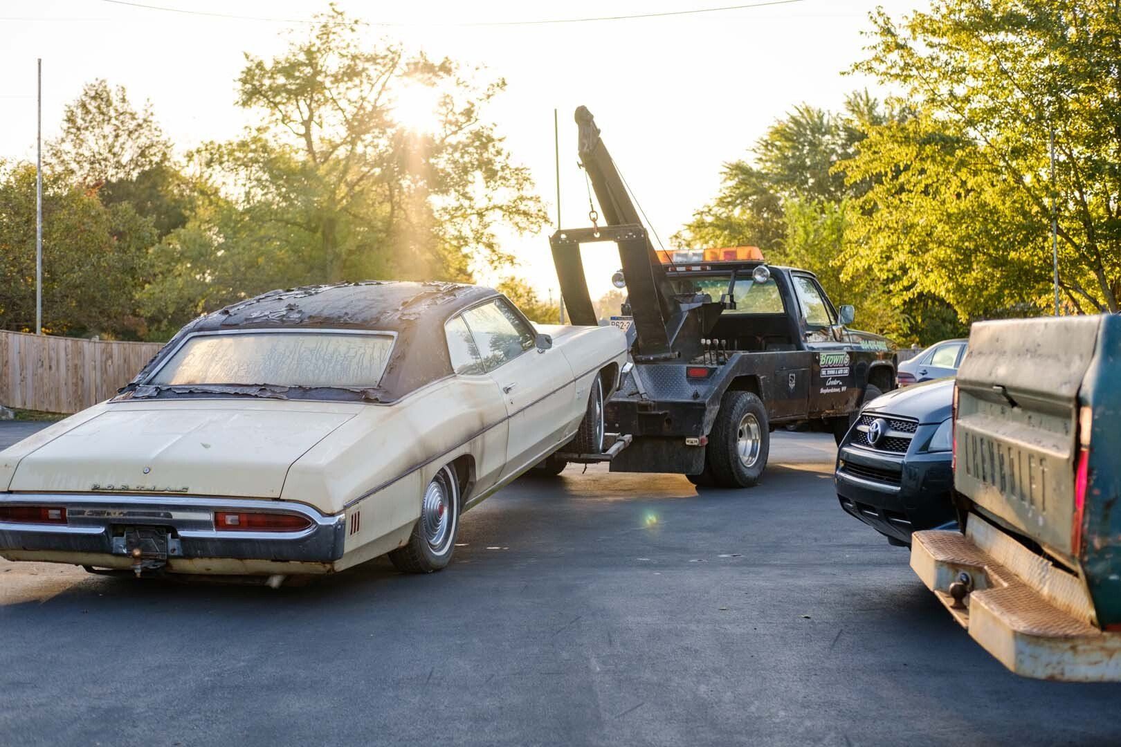 Tow truck lifting a beige classic car in a parking lot. Sunlight shines from behind the vehicles.