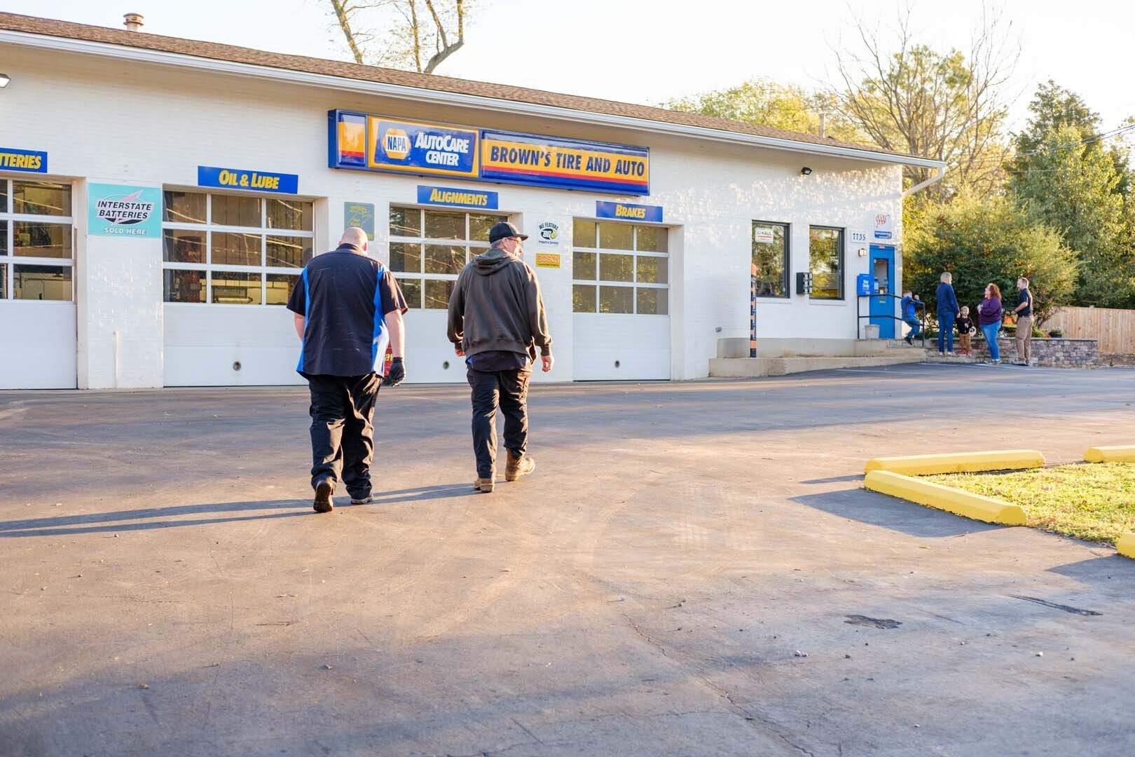 Two people walking away from a car repair shop with garage bays, a Shell sign, and people near a blue gas pump.