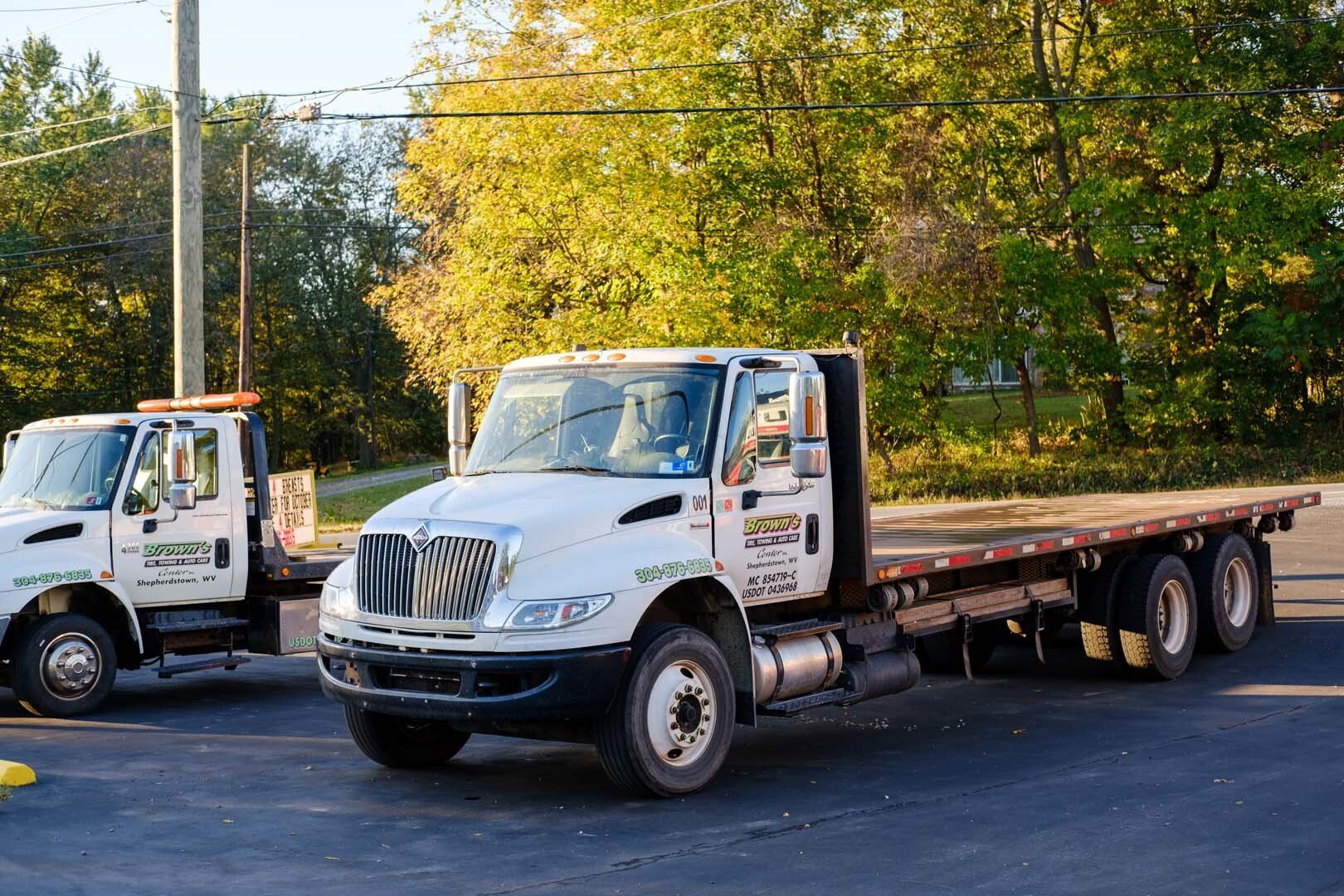 White flatbed truck parked in a lot, another truck visible to the left. Sunny day.