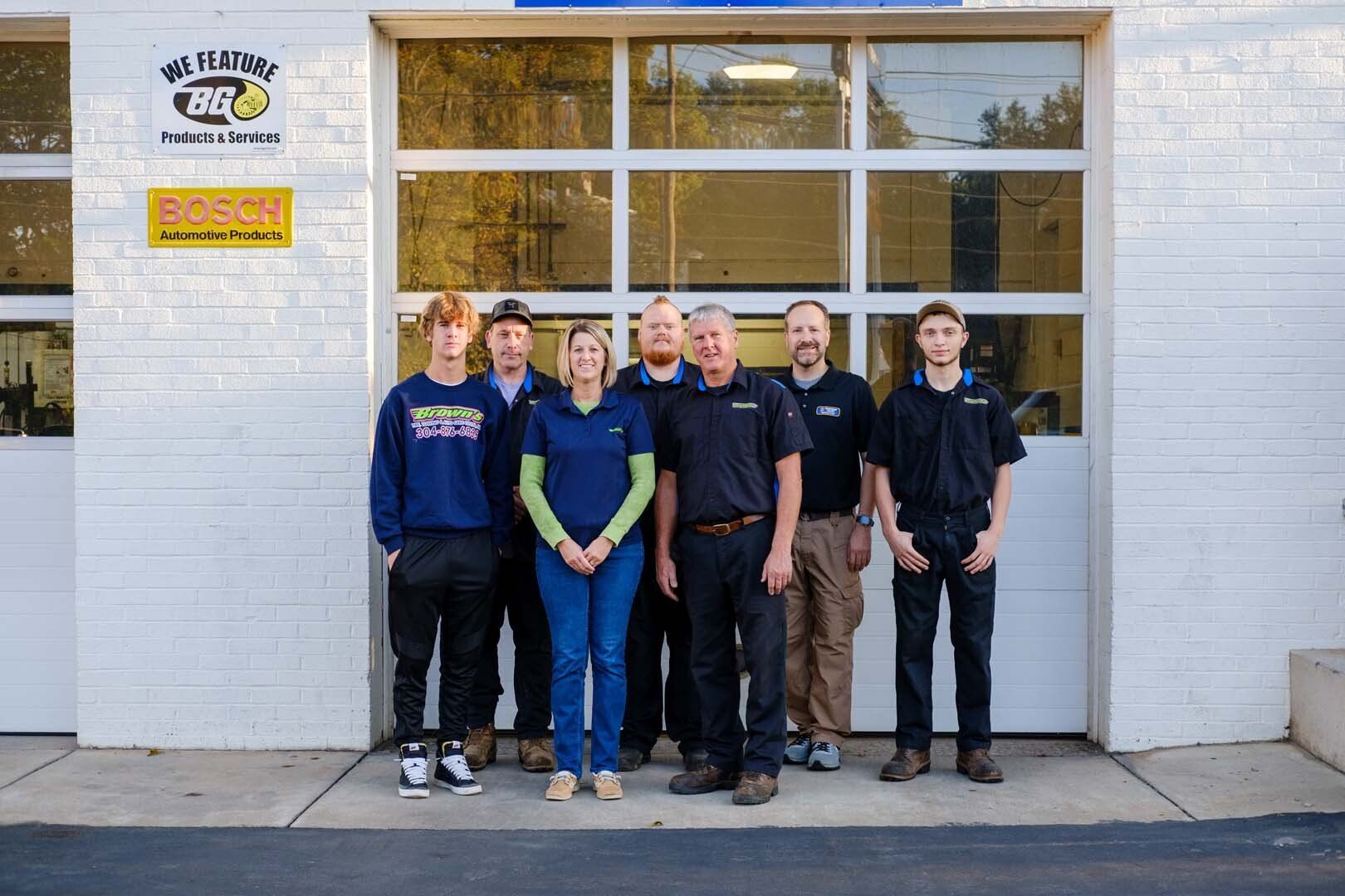 A group of seven people stand in front of a garage door; they are all wearing work uniforms.