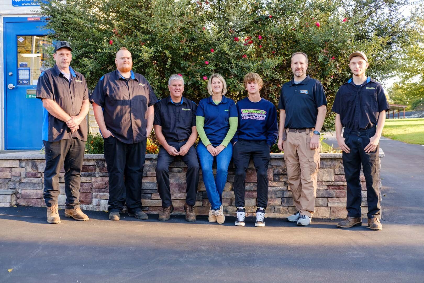 Group of seven people posing in front of a building and foliage; some are wearing work uniforms.