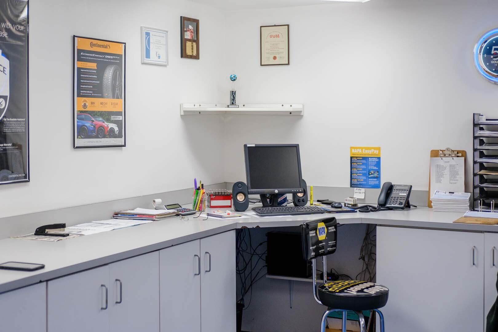 L-shaped office desk in auto shop. Computer, phone, paperwork, and cabinet storage. White walls and fluorescent lighting.