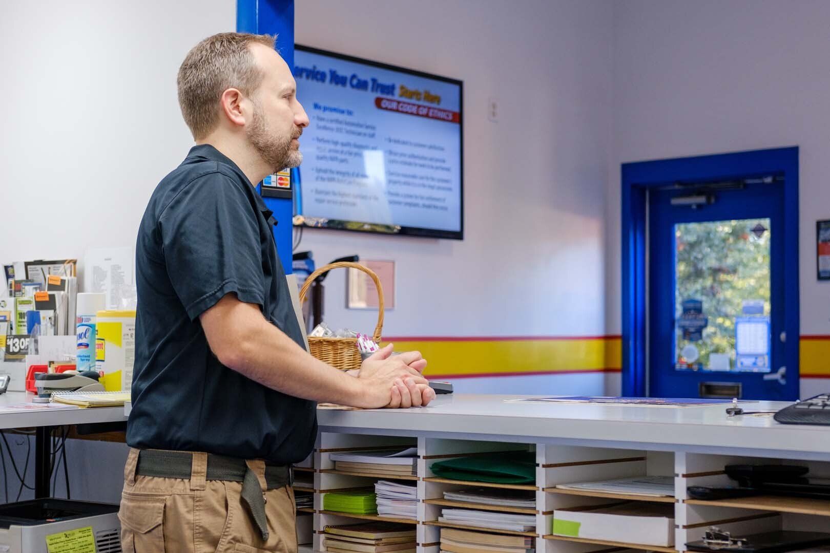 Man stands behind a counter in an auto repair shop, facing a television screen displaying text.